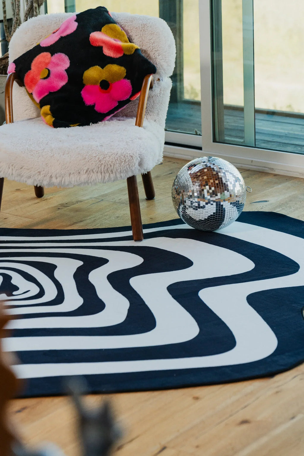 Wavy-patterned rug in black and white paired with a fuzzy chair, floral cushion, and disco ball near a glass sliding door.