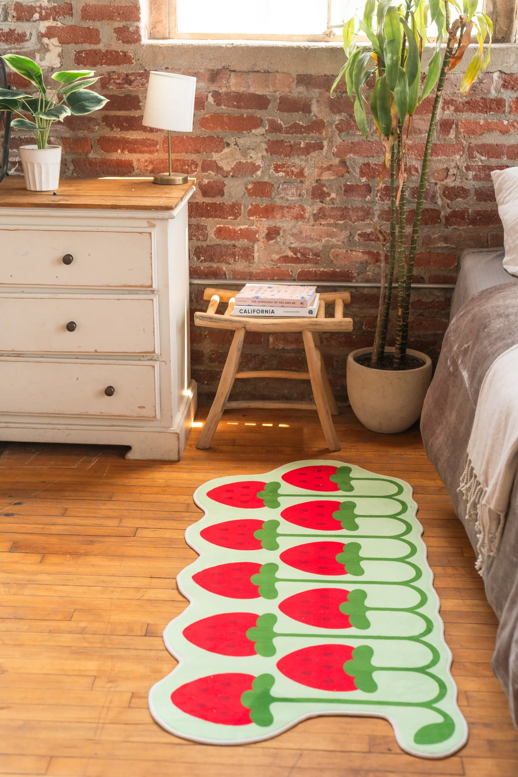 Cute rug with red strawberry design placed beside bed, vintage drawer and houseplants on wooden floor