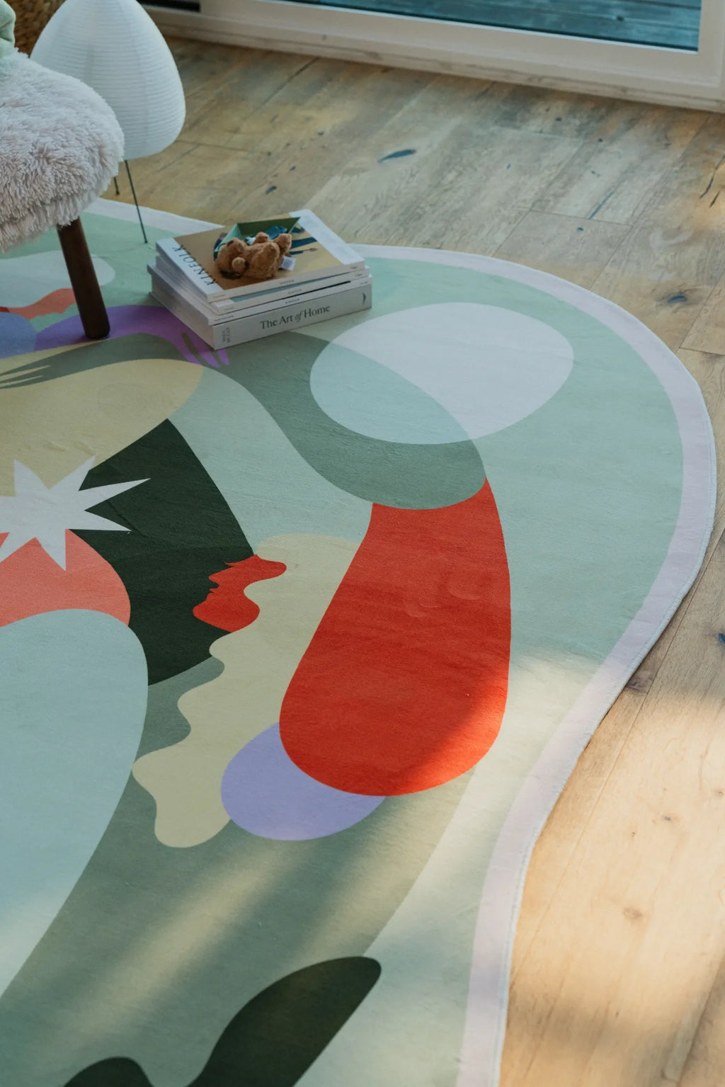 Detail of the rug’s pastel shapes with a stack of books resting on one edge, set in a sunlit room with wooden floor.