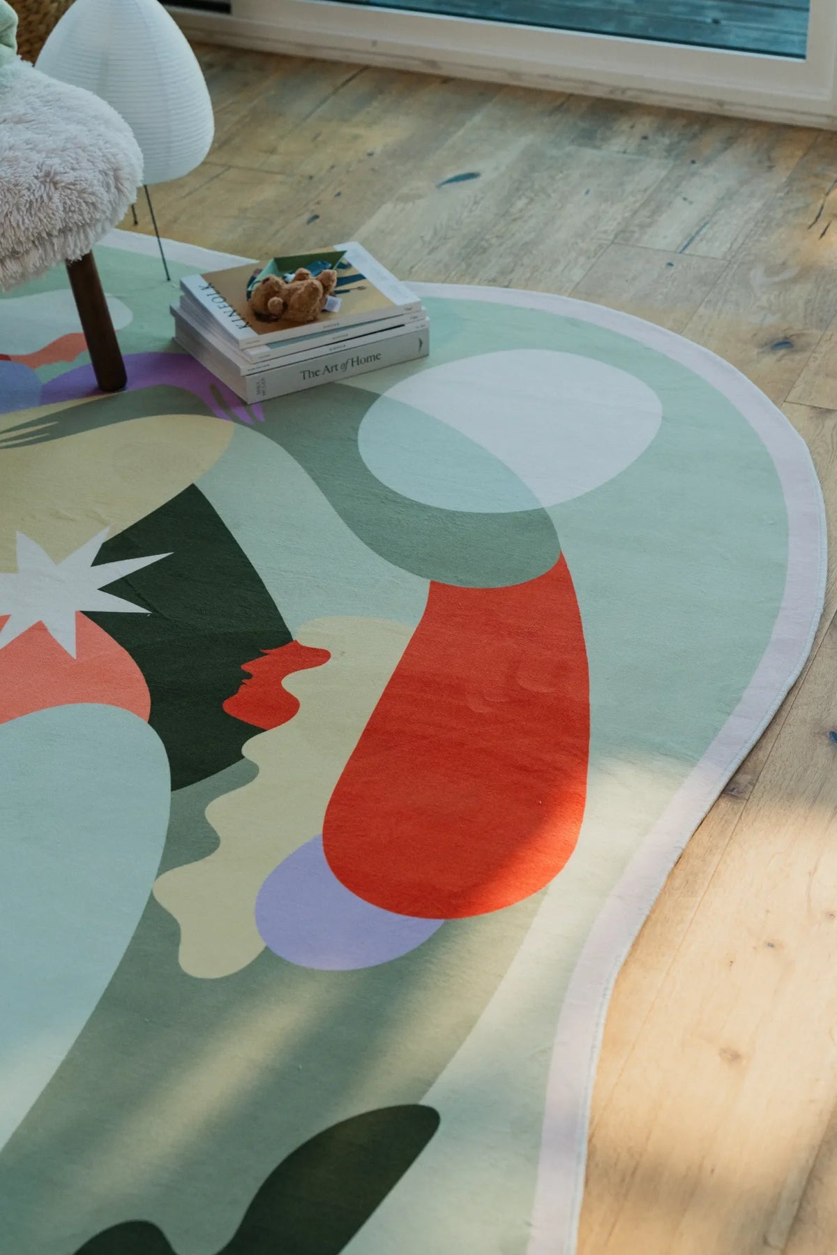 Detail of the rug’s pastel shapes with a stack of books resting on one edge, set in a sunlit room with wooden floor.