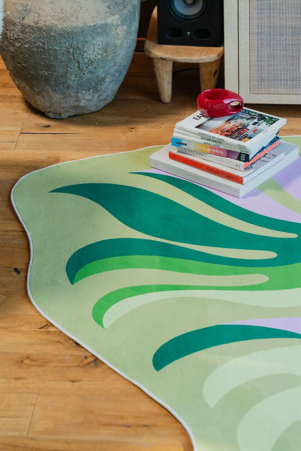 Botanical rug detail next to stacked books and a stone vase on wooden floor, highlighting the wavy green and pastel pattern.