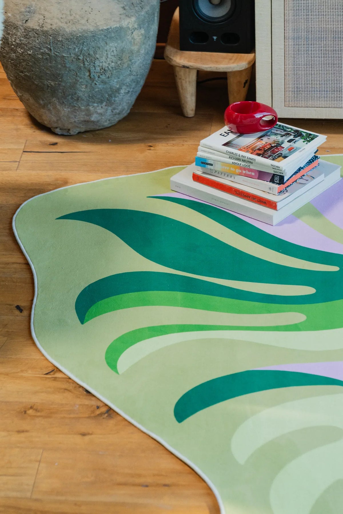 Botanical rug detail next to stacked books and a stone vase on wooden floor, highlighting the wavy green and pastel pattern.
