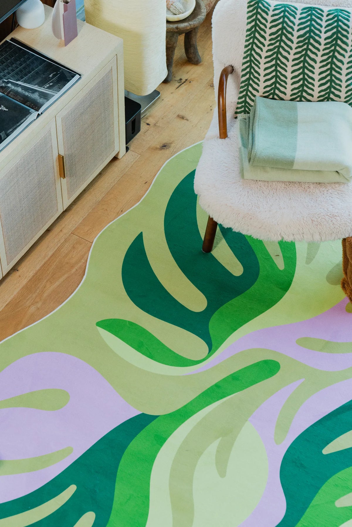 Corner detail of the botanical leaves rug with folded throws on a sherpa chair, soft daylight streaming through the glass door.
