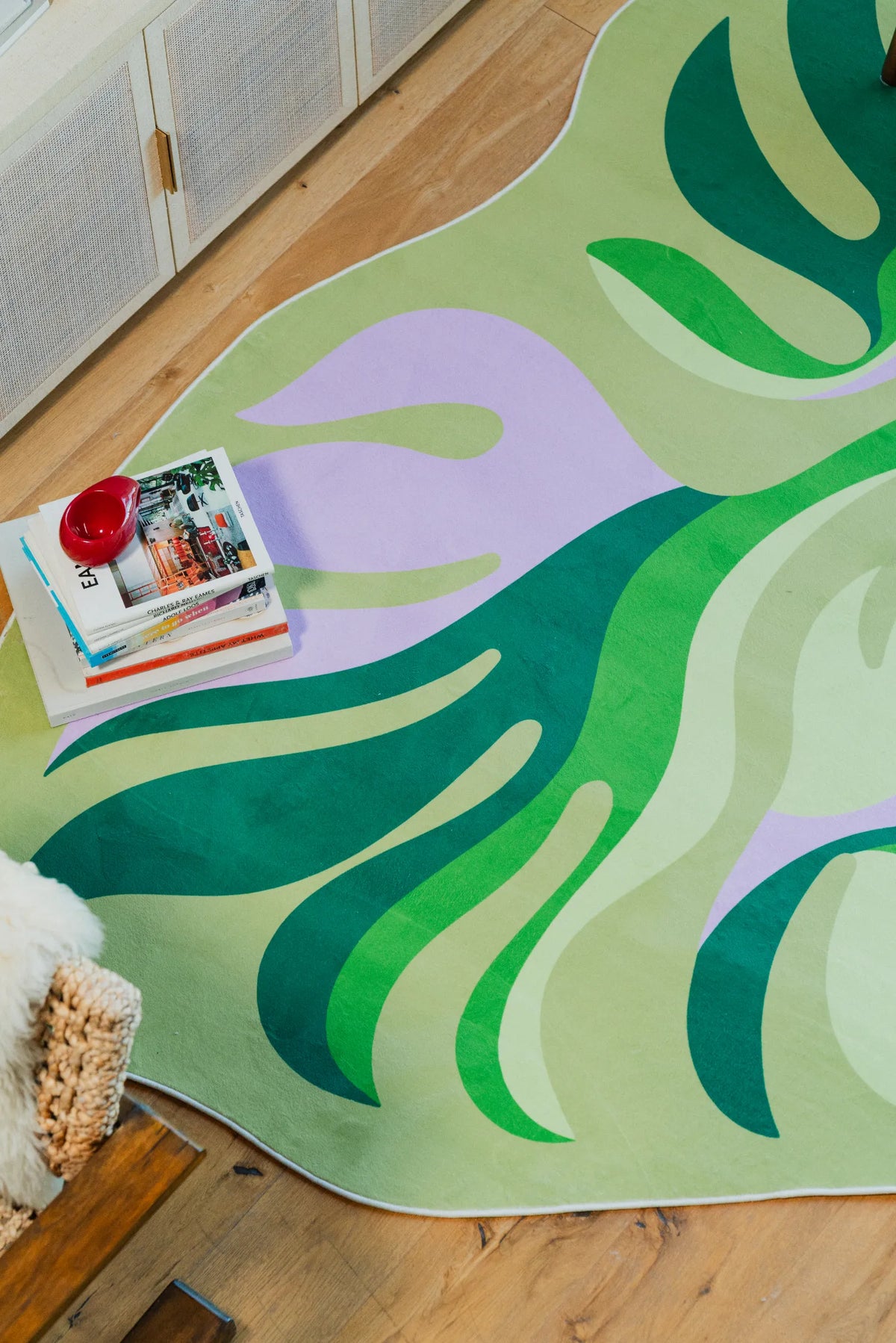 Close-up of the Irregular Botanical Leaves Rug in green, purple, and pink tones, beside a stack of books on wooden floor.