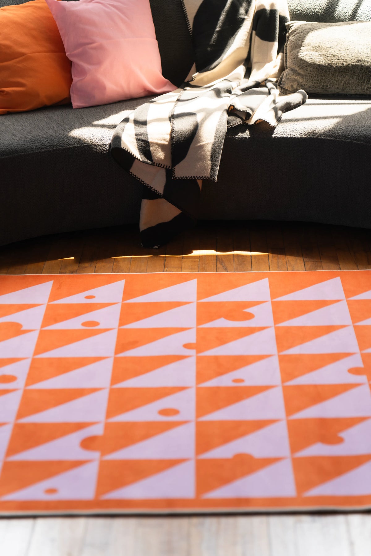Colorful triangle rug in orange and pink tones shown close-up on a wooden floor with sunlight in the background