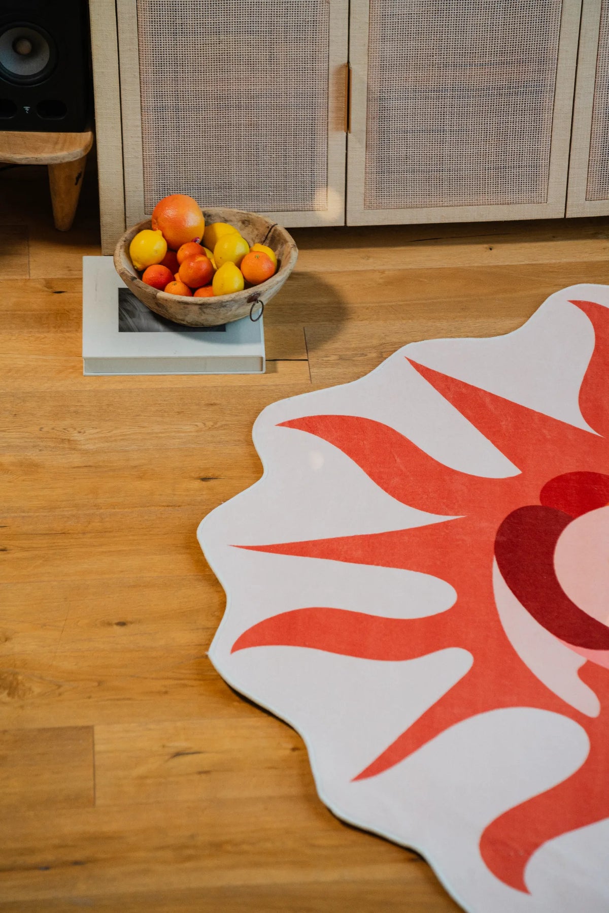 Corner detail of the irregular sun-shaped rug with red and orange rays, next to a woven cabinet and fruit bowl on the floor.