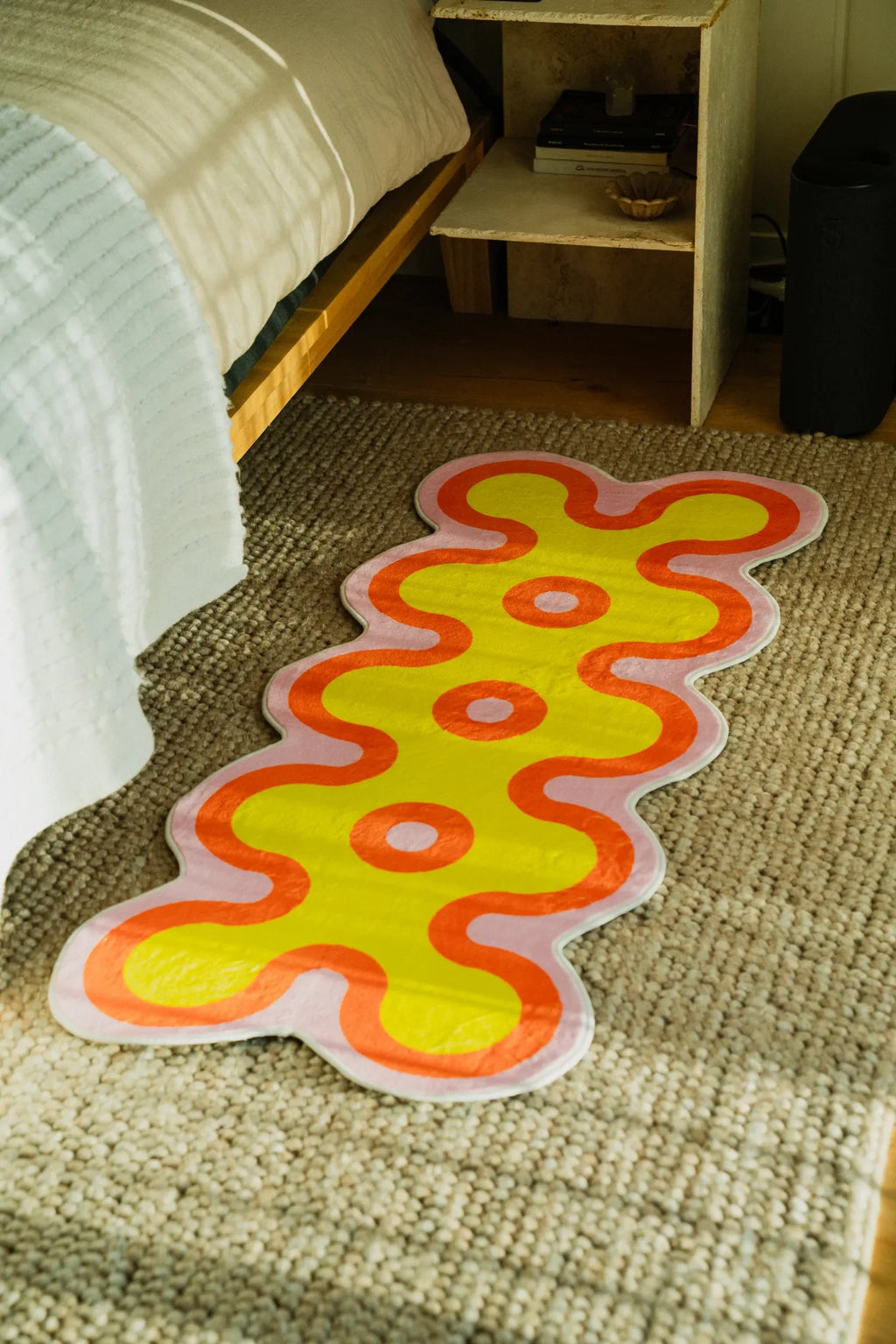 Colorful coral-style runner rug beside a bed on beige carpet, showing playful wavy shapes in yellow and orange tones.