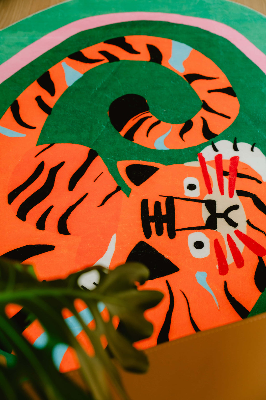 Close-up of the green tiger rug showing its coiled tail and expressive face, partially obscured by indoor plant leaves.
