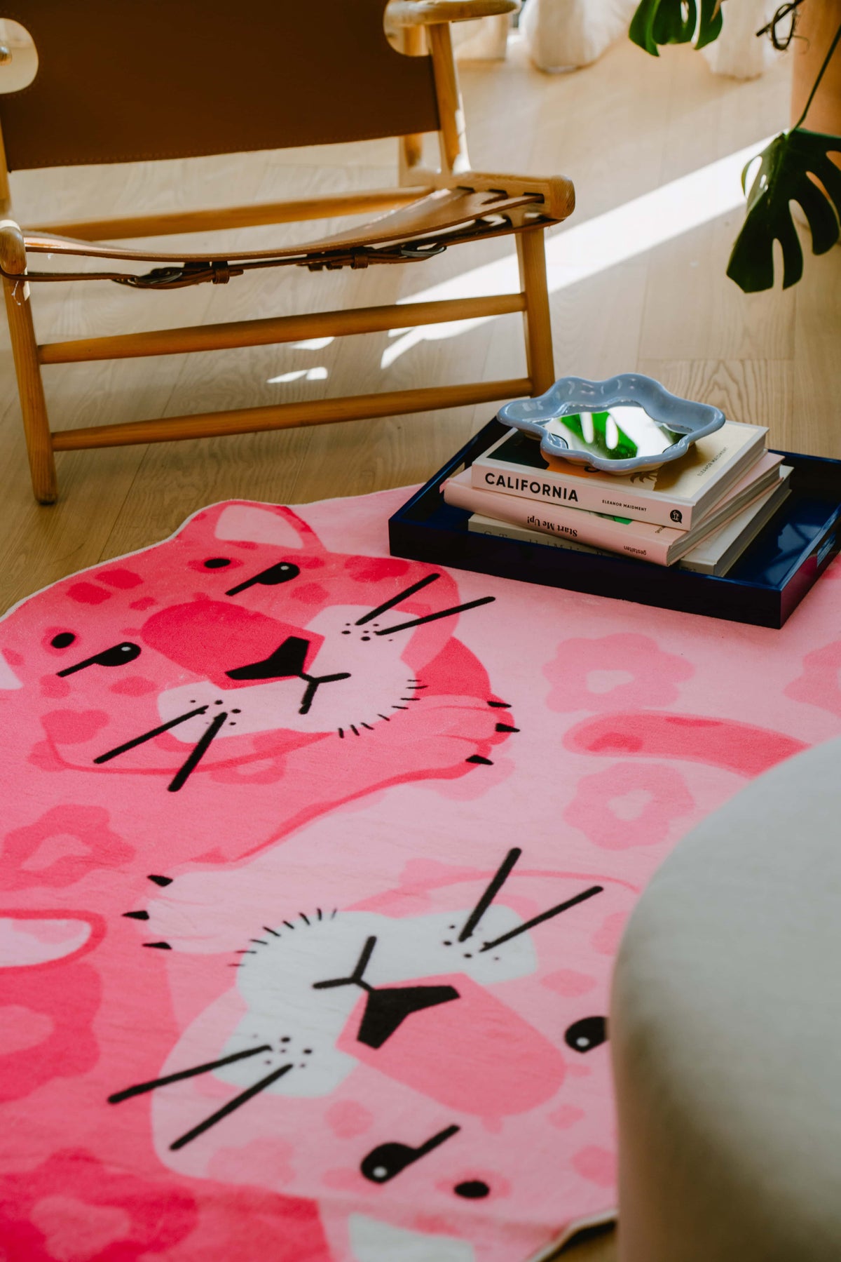 Two pink panthers illustrated on the rug with books and greenery around, placed beside a wooden lounge chair in sunlit nook.