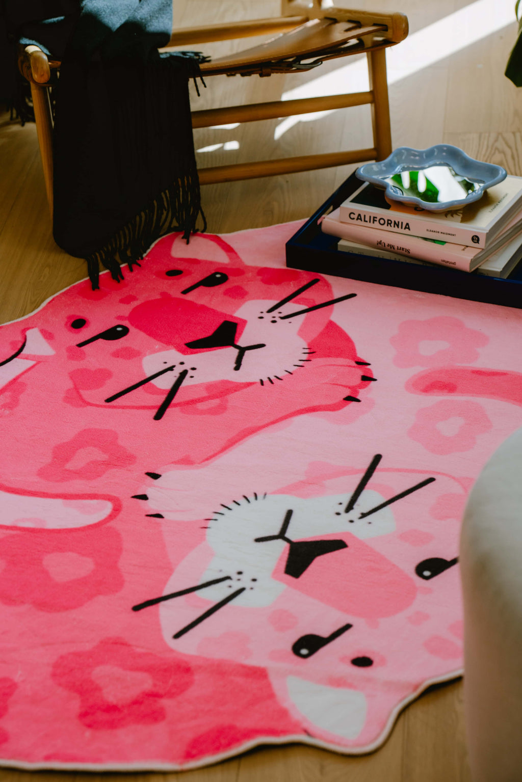 Pink panther rug with floral print laid in front of a wooden lounge chair, styled with black throw and books on a tray.